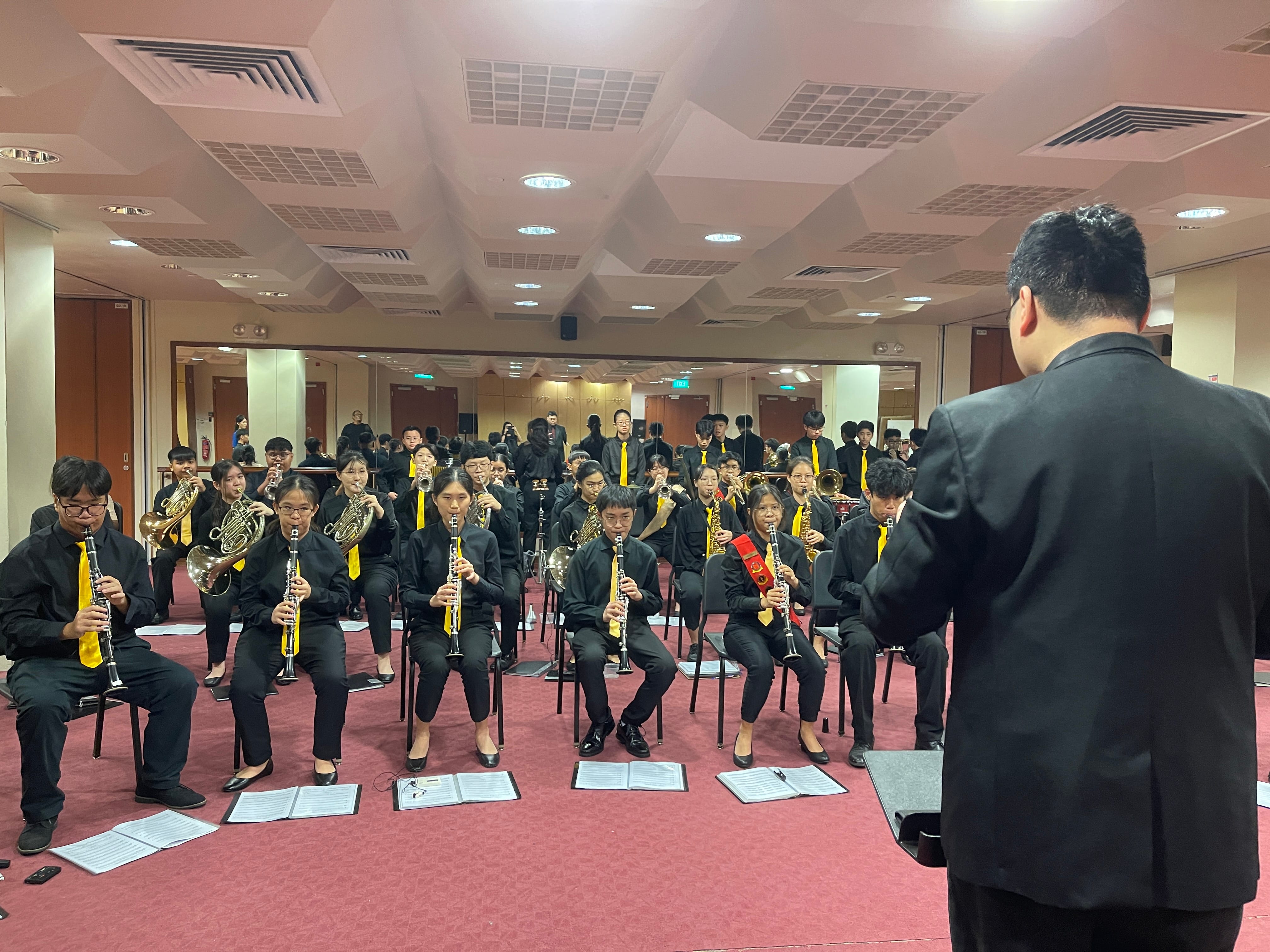 A conductor in a black suit leads a youth wind band wearing black uniforms with yellow ties in an indoor hall.